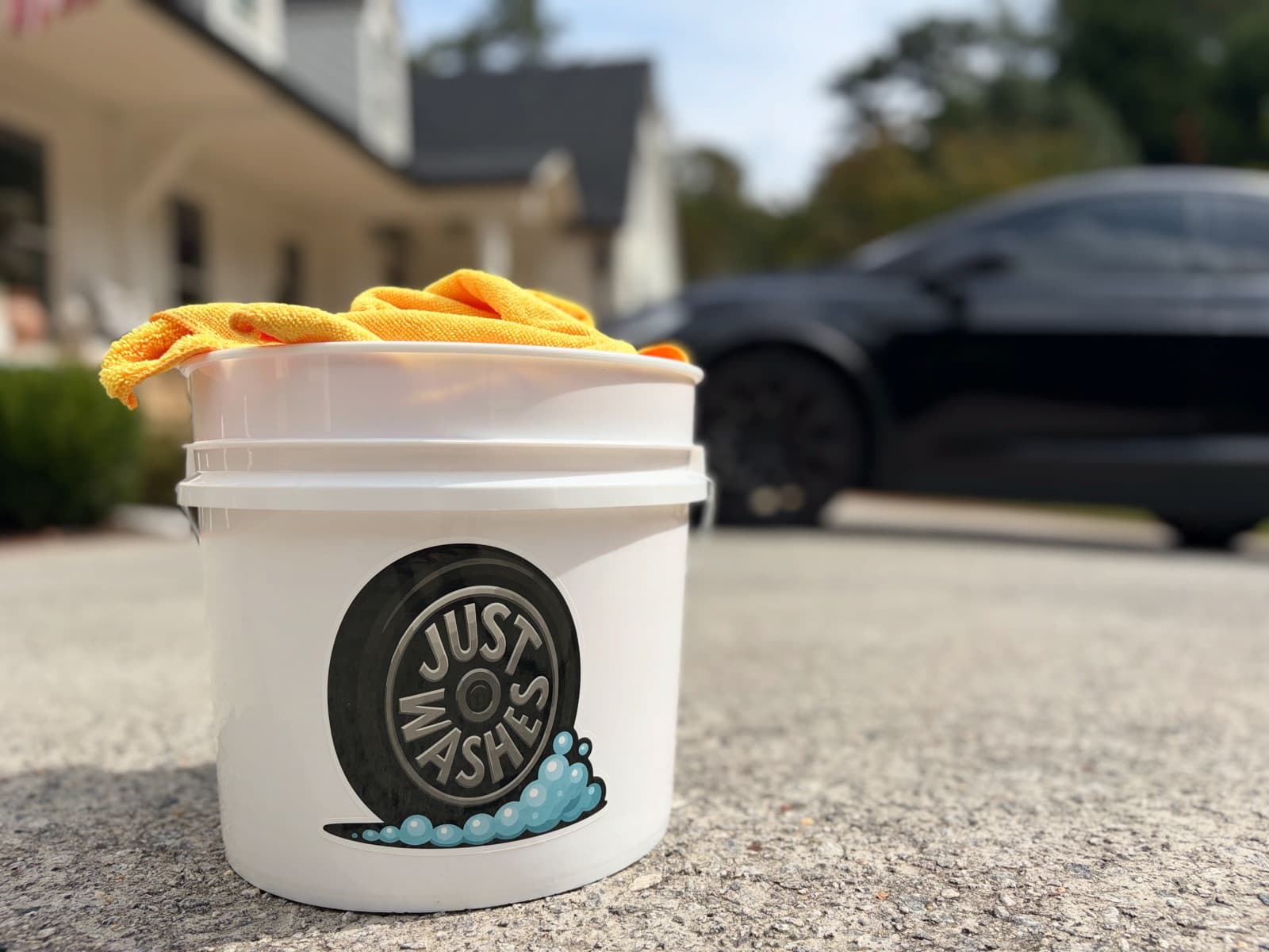 JustWashes bucket with yellow microfiber towels on a customer's driveway, a dark car behind it